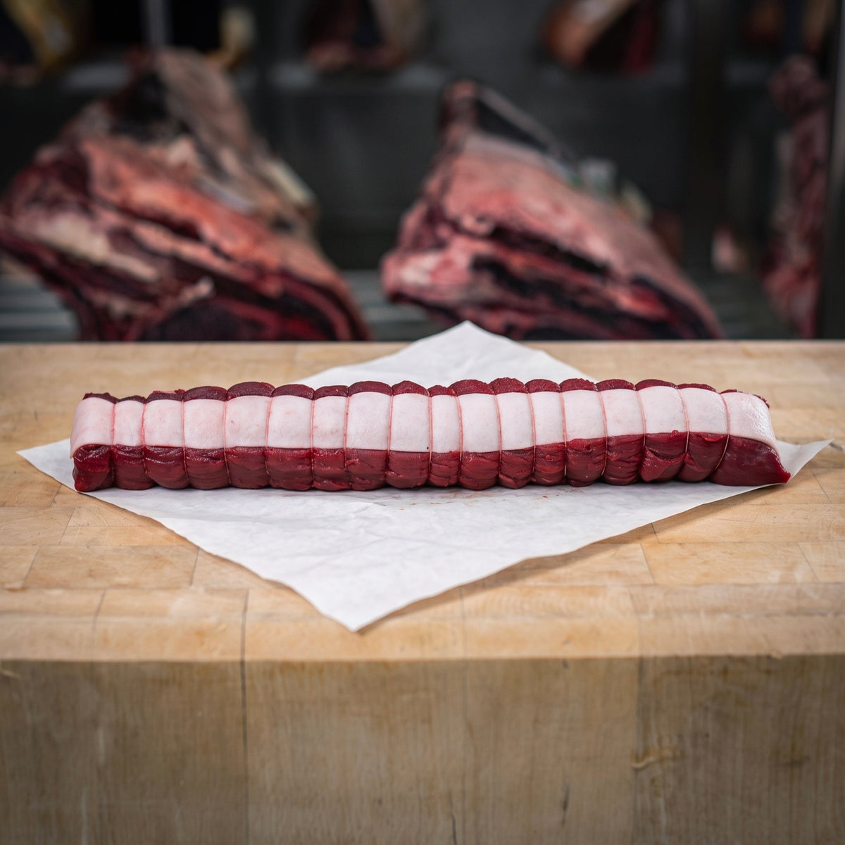 Filet de biche et cerf cru d’environ 500 g, paré, bardé de lard de porc et ficelé, sur un billot en bois